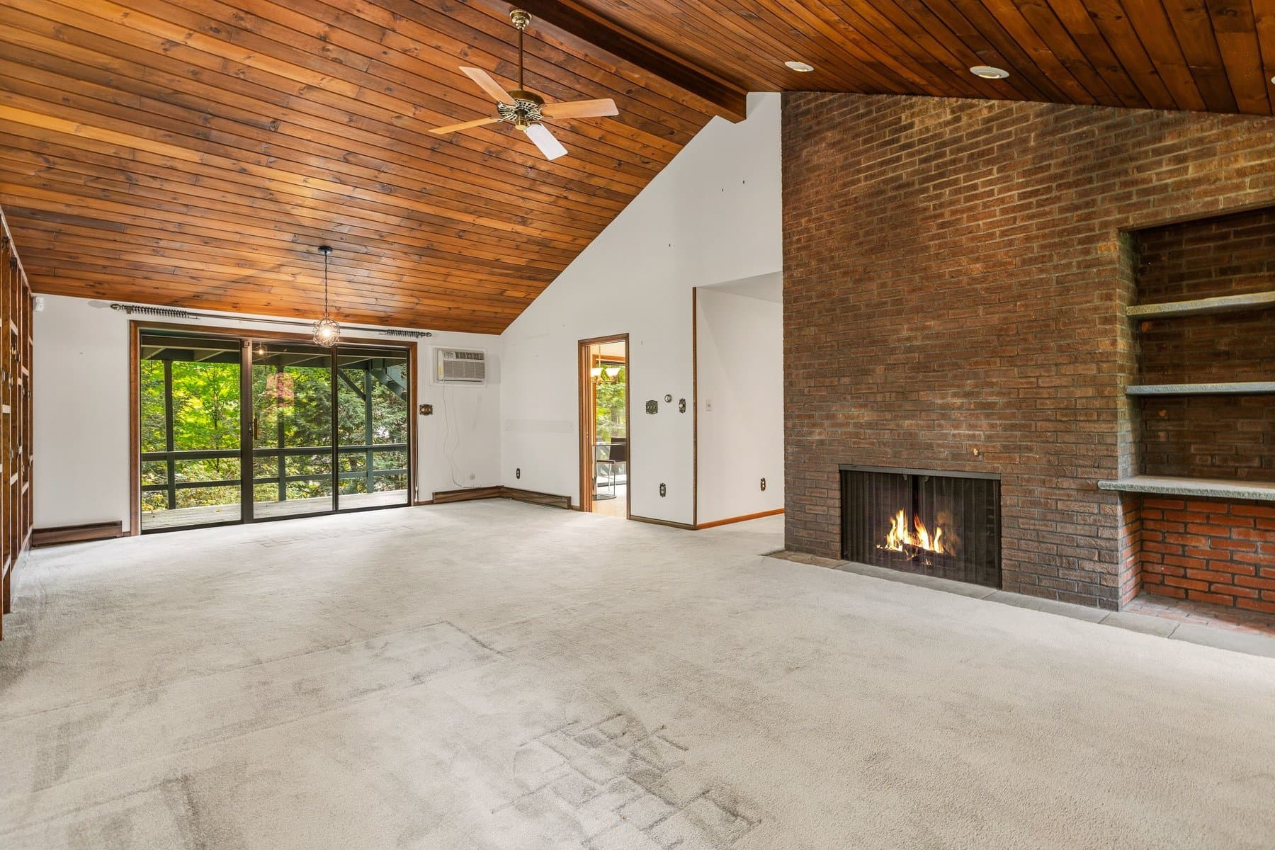 The living room under the vaulted wood-plank ceiling, with the brick fireplace lit and the wall of windows to the woods